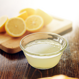 A small bowl of lemon juice sits on a wooden table in front of some sliced lemons. 