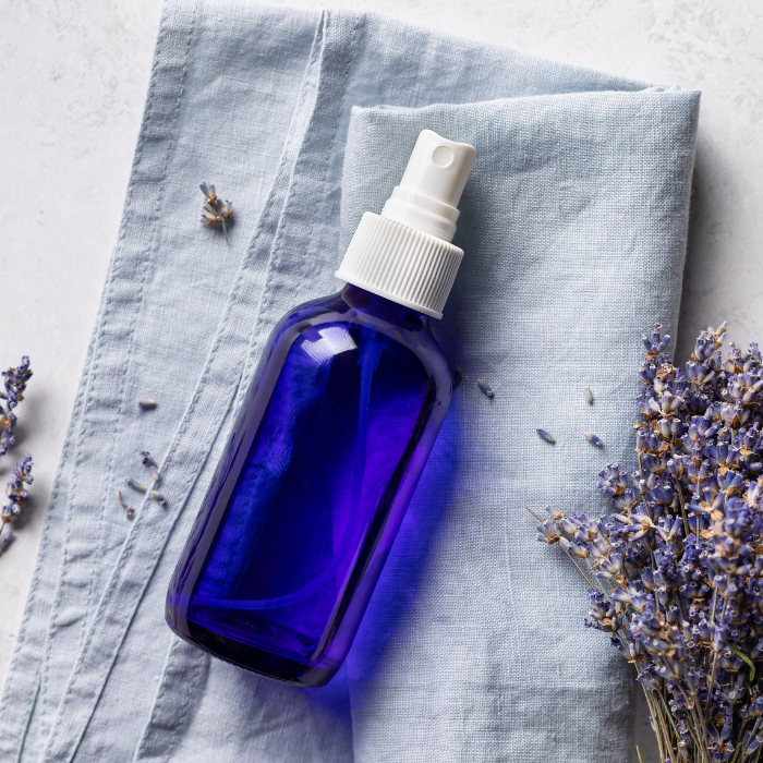 Lavender hydrosol in a glass sprayer bottle next to a sprig of lavender on a blue cloth