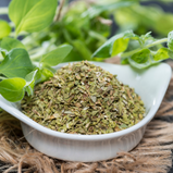 A small white dish filled with loose dried Oregano sits on a background of fresh oregano,