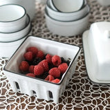 A white porcelain berry basket shown here as part of a tabletop scene and containing blackberries and raspberries.