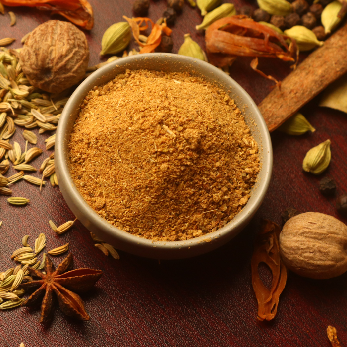 Garam masala in a ceramic bowl surrounded by whole spices on a wooden background